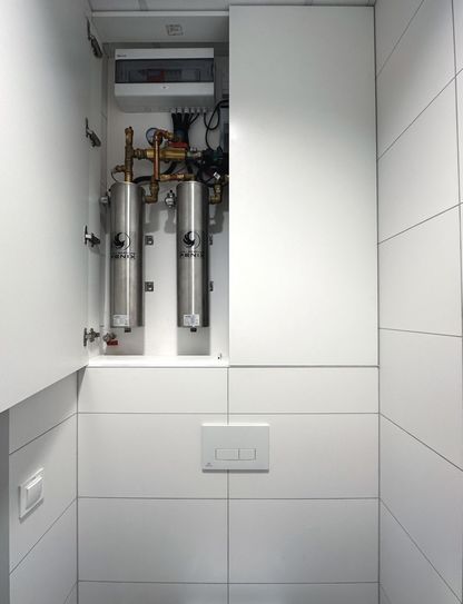 Instalacja 9 kW w białej obudowie w łazience.Szafka jest do połowy zamknięta. Pod instalacja jest spłuczka toaletowa.
